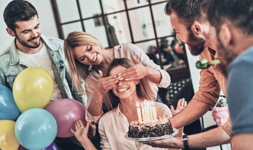 Četri draugi apsveic sievieti dzimšanas dienā ar garšīgu kūku un daudz baloniem.